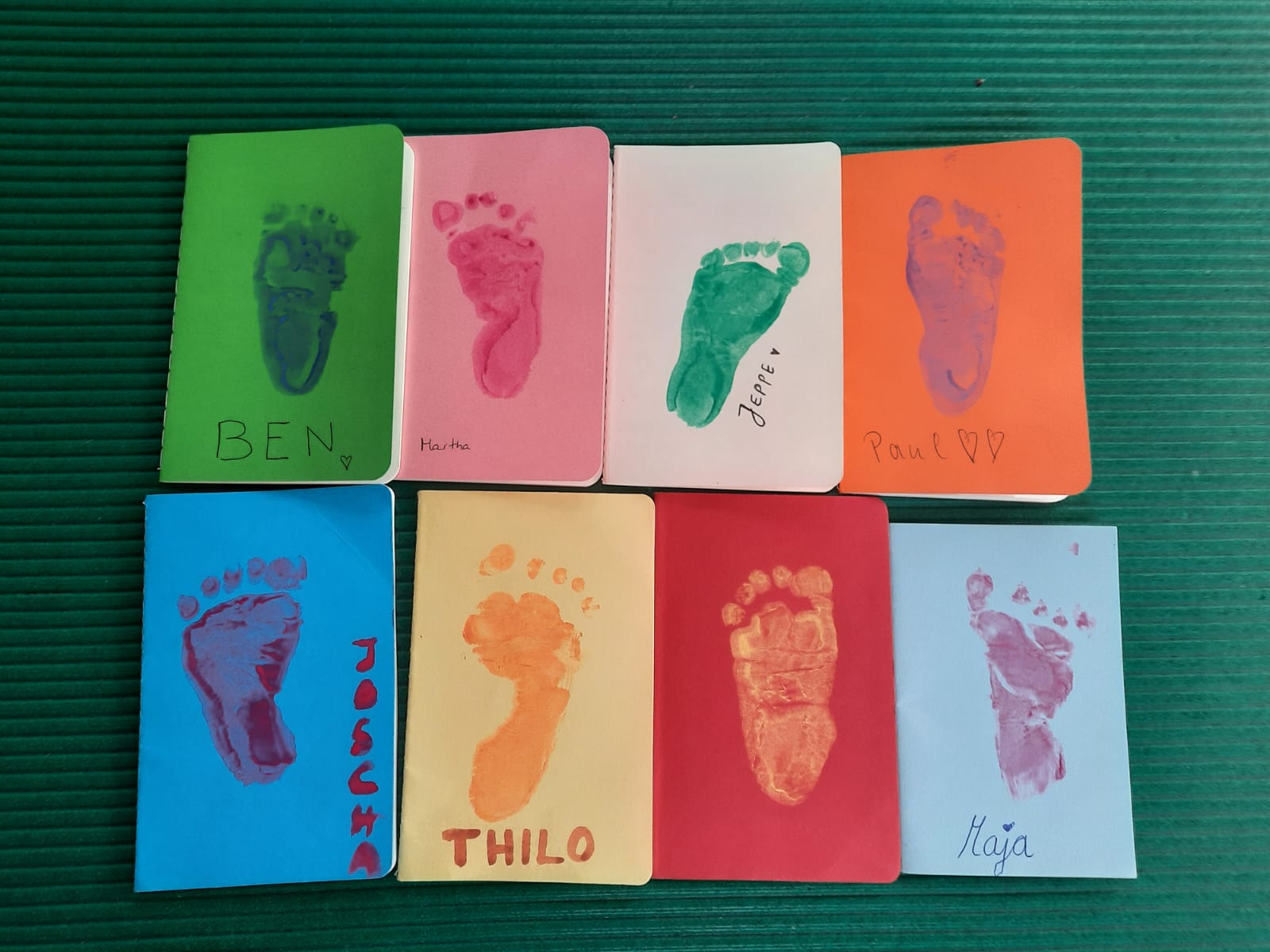 Vier bunte Blätter, auf denen jeweils ein farbiger Fußabdruck von einem Kind zu sehen ist. Darunter steht der Name des Kindes. Die Bilder sind im Rahmen einer der Eltern-Kind-Kurse entstanden..
